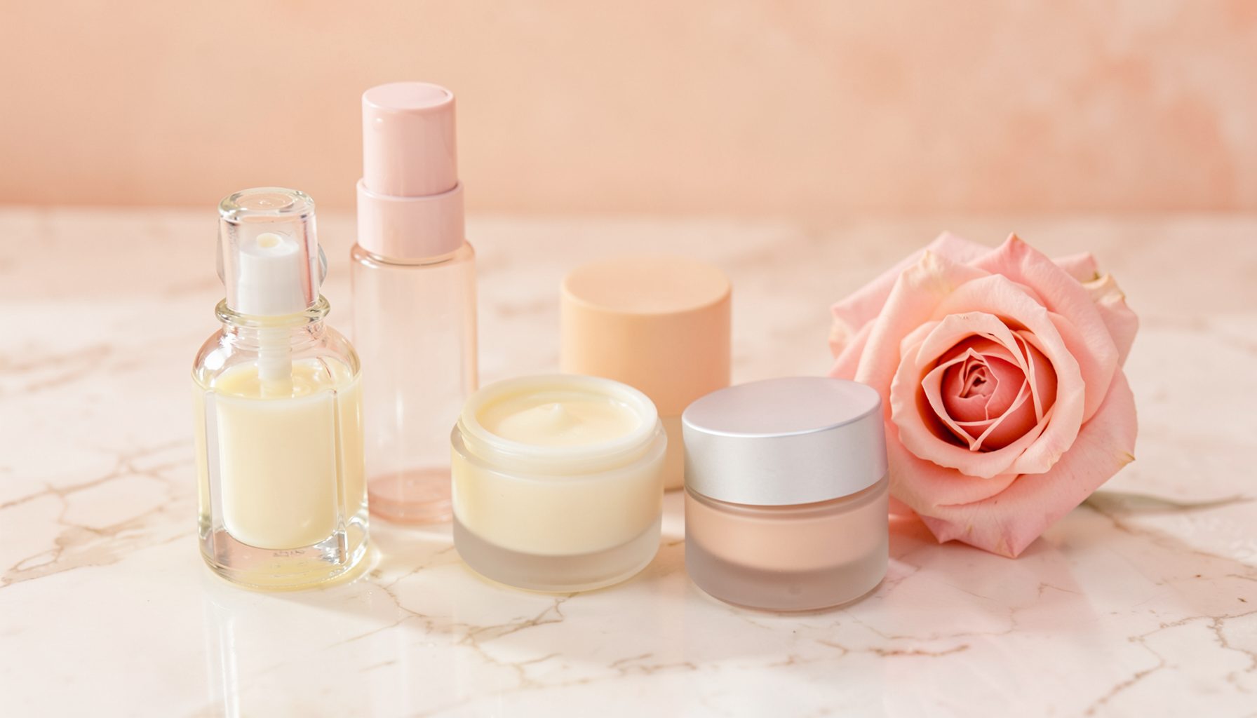 An assortment of beauty and skincare products — glass bottles, cream jars, and a soft pink rose — arranged on a marble surface in warm pastel tones.