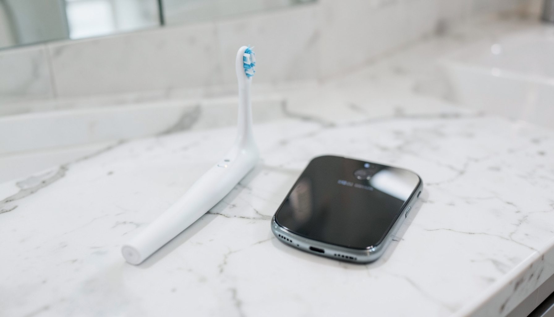 A sleek electric toothbrush and a smartphone side by side on a marble counter, clean modern bathroom aesthetic.